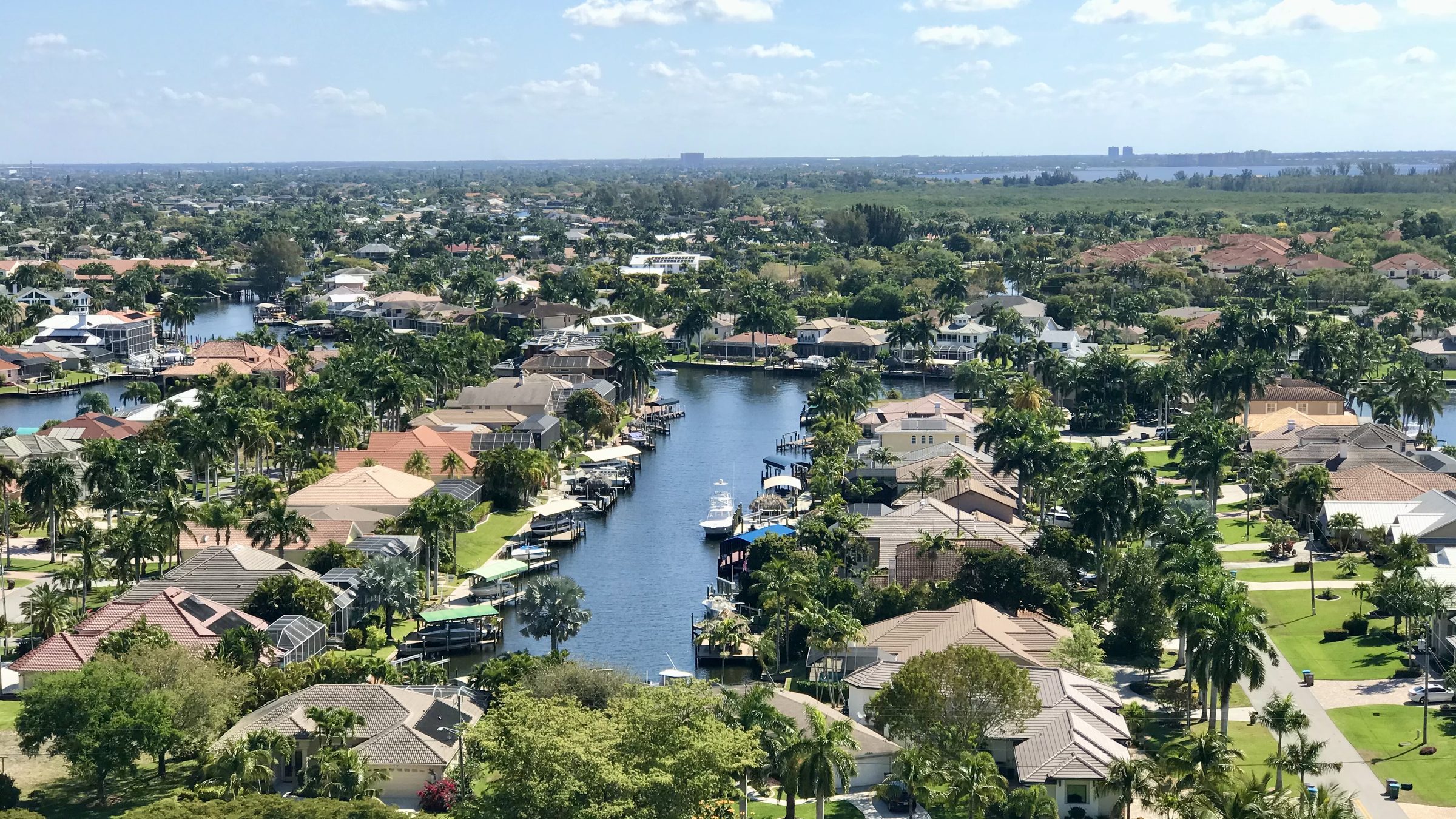 Looking down a Cape Coral residential canal from above: docked boats, screened lanais, tile roofs and palms running out toward the river.