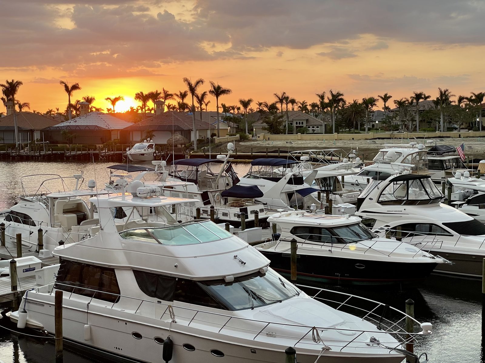 Sunset at Cape Harbour marina: motor yachts moored in their slips, the sun dropping behind a row of palms and tile-roofed houses across the basin.